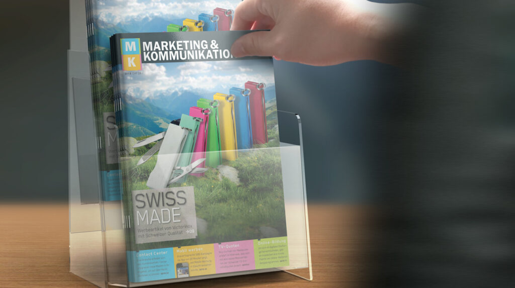 Eine Hand greift aus einem durchsichtigen Acryldisplay nach einem Magazin mit dem Titel „Marketing & Kommunikation“. Das Cover zeigt farbenfrohe Mappen vor einer malerischen Bergkulisse. Auf dem Cover steht unter anderem „Swiss Made“. Das Display ist auf einer Holzoberfläche platziert. von Clicdesign AG, Werbung und Webdesign
