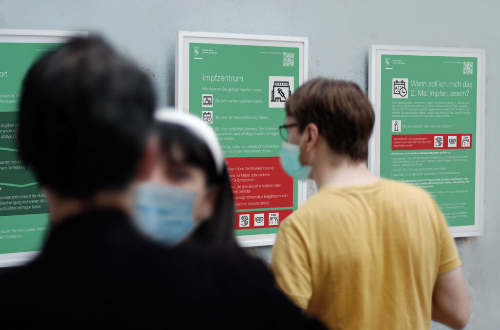 Menschen betrachten Informationsplakate über Impfungen in deutscher Sprache, die von Clicdesign AG gestaltet wurden. Die Plakate mit Texten und Symbolen zu Covid-19-Impfzentren hängen prominent an einer Wand. Als Teil des Projekts für einen Kunden aus dem Gesundheitswesen veranschaulichen diese Aufklärungsgrafiken die Kompetenz von Clicdesign AG in den Bereichen Werbung und Printdesign. Einige Personen tragen Masken, während sie sich die visuell ansprechenden Grafiken ansehen, was das Engagement von Clicdesign AG für effektive Kommunikation im Webdesign und in Printmedien innerhalb der Werbebranche unterstreicht.